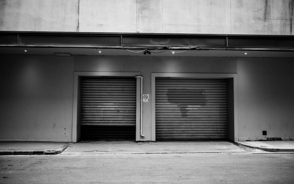 A monochrome urban exterior with two closed garage doors and a concrete wall.